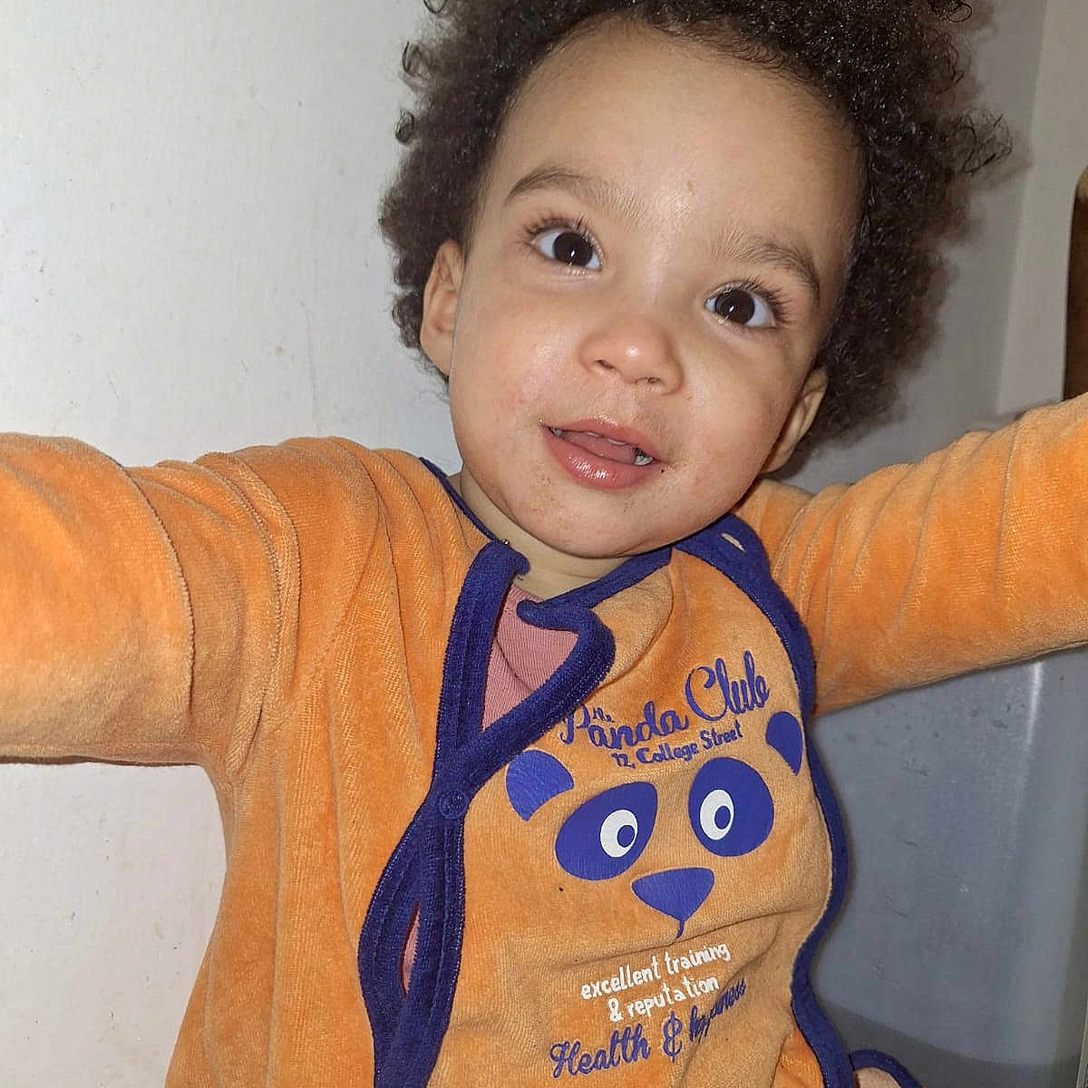 Ami a rejoint le concours — aidez-le/la à gagner de superbes lots ! bin, blue_trim, casual, child, curly_hair, cute, eyes, face, indoor, long_sleeves, mouth, orange_clothing, panda_design, person, portrait, reaching, sitting, toddler, wall, young_child