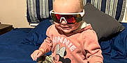 Amelia is registered to the contest to win money with this photo: baby, child, sunglasses, pink_sweatshirt, minnie_mouse, camouflage_pants, bed, pillow, striped_curtain, pacifier, foot, hand, indoor, smile, clothing, person, cute, fashion, accessory, toddler