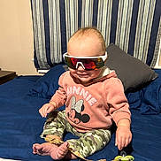 Amelia joined the competition — help win amazing prizes! baby, child, sunglasses, sweatshirt, minnie_mouse, camouflage_pants, bed, pillow, blanket, indoor, smiling, person, foot, hand, toy, pacifier_clip, curtains, striped, casual_clothing, cute
