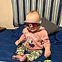 baby, child, sunglasses, sweatshirt, minnie_mouse, camouflage_pants, bed, pillow, blanket, indoor, smiling, person, foot, hand, toy, pacifier_clip, curtains, striped, casual_clothing, cute