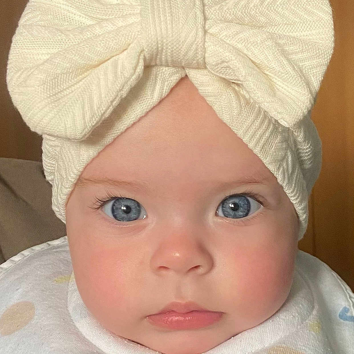 Amelia is registered to the contest to win money with this photo: baby, infant, face, blue_eyes, knitted_hat, bow, rosy_cheeks, close_up, portrait, cute, child, soft_lighting, white_clothing, pastel_colors, expression, indoors, headwear, young_child, skin, eyelashes