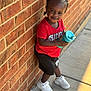 toddler, child, smiling, braided_hair, red_tshirt, black_shorts, white_sneakers, sippy_cup, brick_wall, sidewalk, outdoor, sunlight, shadow, happy, casual_clothing, person, portrait, daylight, urban, leaning