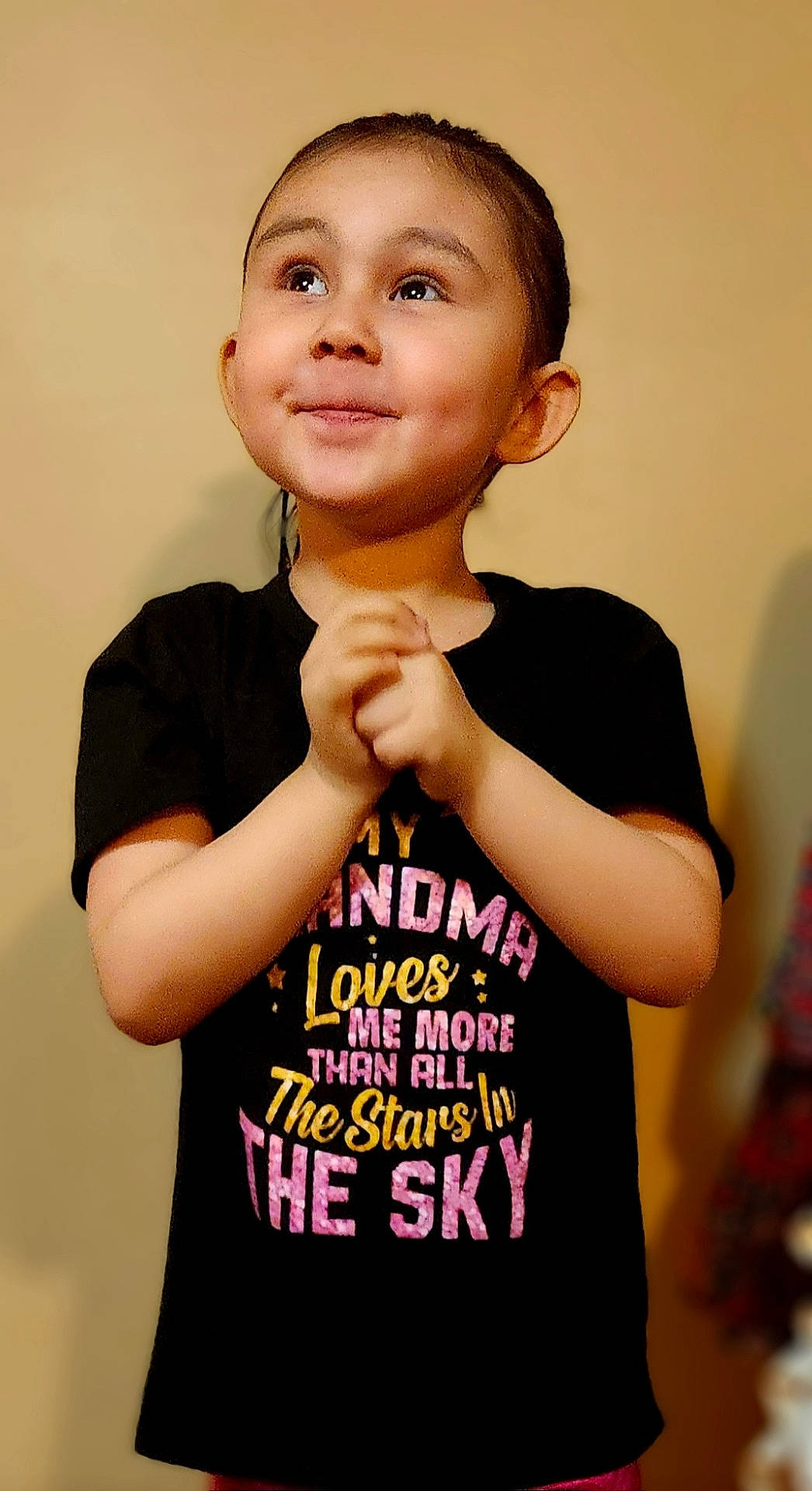 Emma is registered to the contest to win money with this photo: background_blur, black_tshirt, casual_clothing, child, clasped_hands, colorful_text, cute, expression, happy, hopeful, indoor, light_skin, looking_up, person, portrait, short_hair, smiling, text, tshirt, young