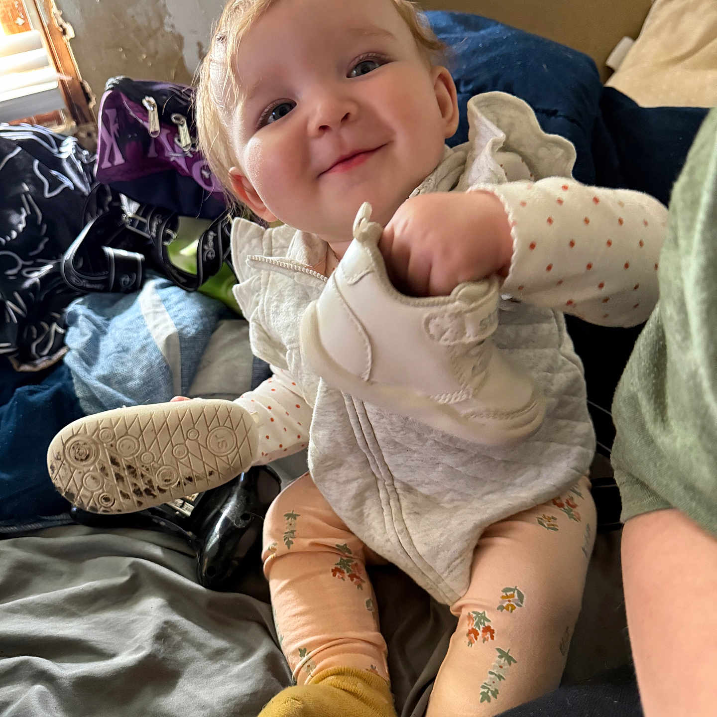 Aurora is registered to the contest to win money with this photo: baby, bed, child, clothing, cushion, cute, face, happy, holding, indoor, light, patterned_pants, person, portrait, shoe, sitting, smiling, white_jacket, window, yellow_socks