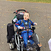 Raven joined the competition — help win amazing prizes! child, wheelchair, costume, captain_america, outdoor, paved_path, autumn_leaves, park, trees, car, person, clothing, footwear, sitting, straps, expression, daylight, wheel, sidewalk, accessory