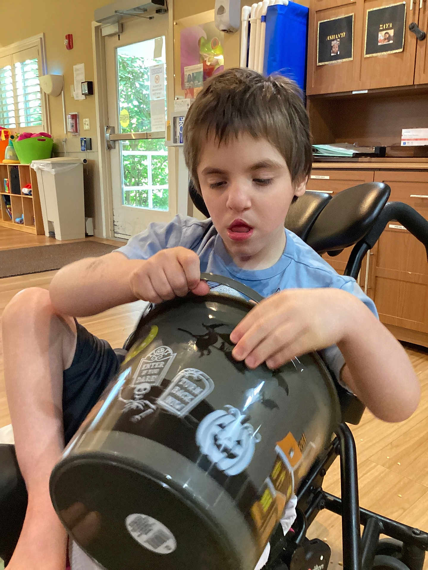 Raven joined the competition — help win amazing prizes! child, boy, wheelchair, indoor, bucket, halloween, pumpkin, tombstone, decorations, wooden_floor, cabinet, light, window, door, shorts, blue_shirt, furniture, person, curtains, plastic_bucket