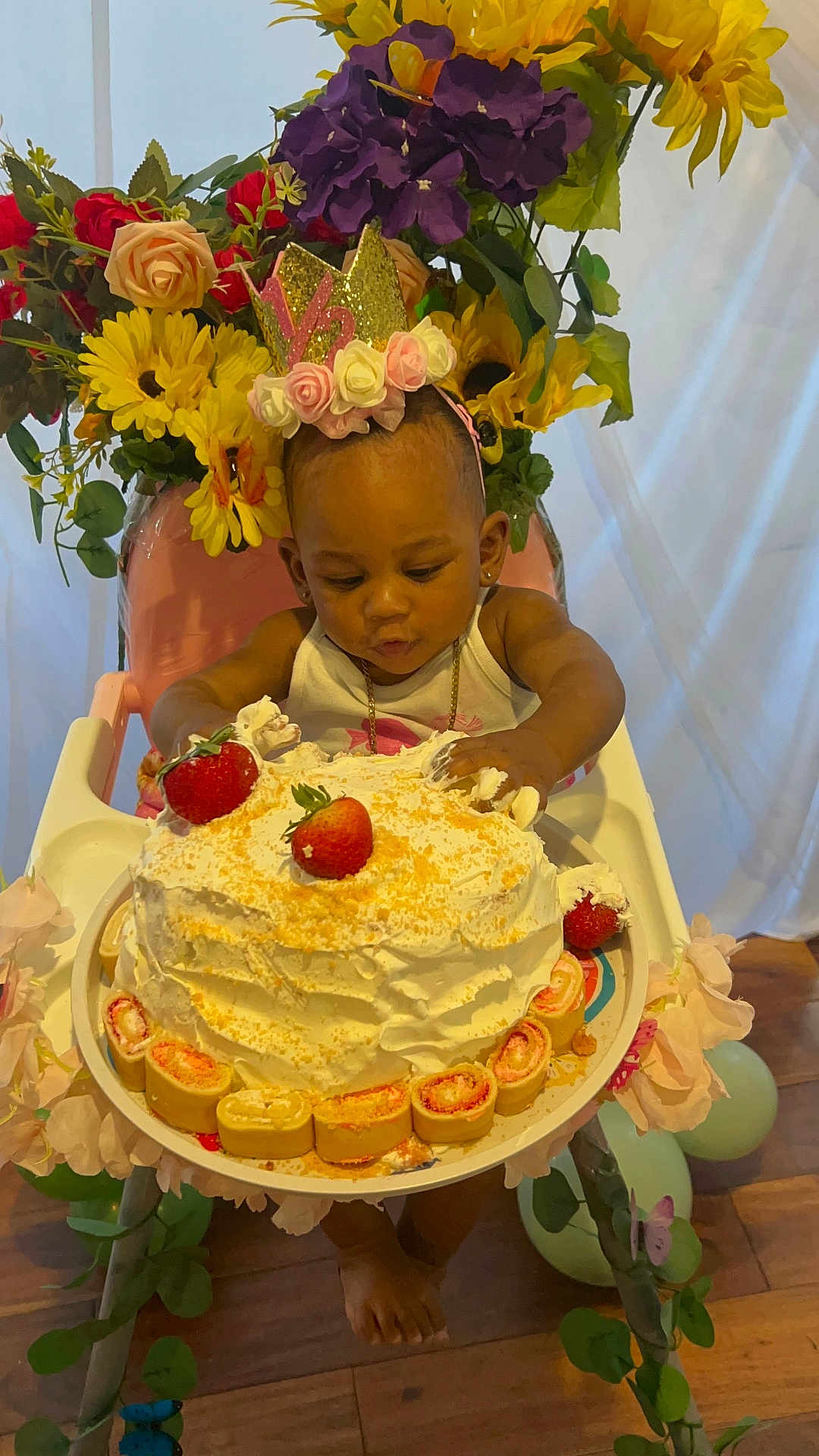 Ailai is registered to the contest to win money with this photo: baby, child, cake, strawberries, high_chair, flower_crown, flowers, cream, birthday, celebration, decorations, curious, cute, indoor, plate, wooden_floor, pink_chair, gold_chain, hand, foot