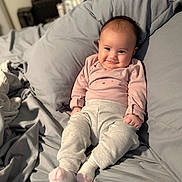 Zuri is registered to the contest to win money with this photo: baby, infant, smiling, bed, pillow, gray_bedding, pink_clothing, cozy, indoors, cute, child, portrait, soft_lighting, socks, relaxed, young, person, candid, happy, sleepwear