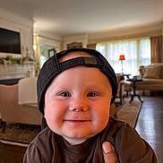 Canaan is registered to the contest to win money with this photo: baby, child, smiling, cap, indoor, living_room, furniture, hand, person, cute, portrait, brown_shirt, cozy, cheeks, headwear, happy, home, toddler, face, casual