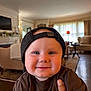 baby, child, smiling, cap, indoor, living_room, furniture, hand, person, cute, portrait, brown_shirt, cozy, cheeks, headwear, happy, home, toddler, face, casual