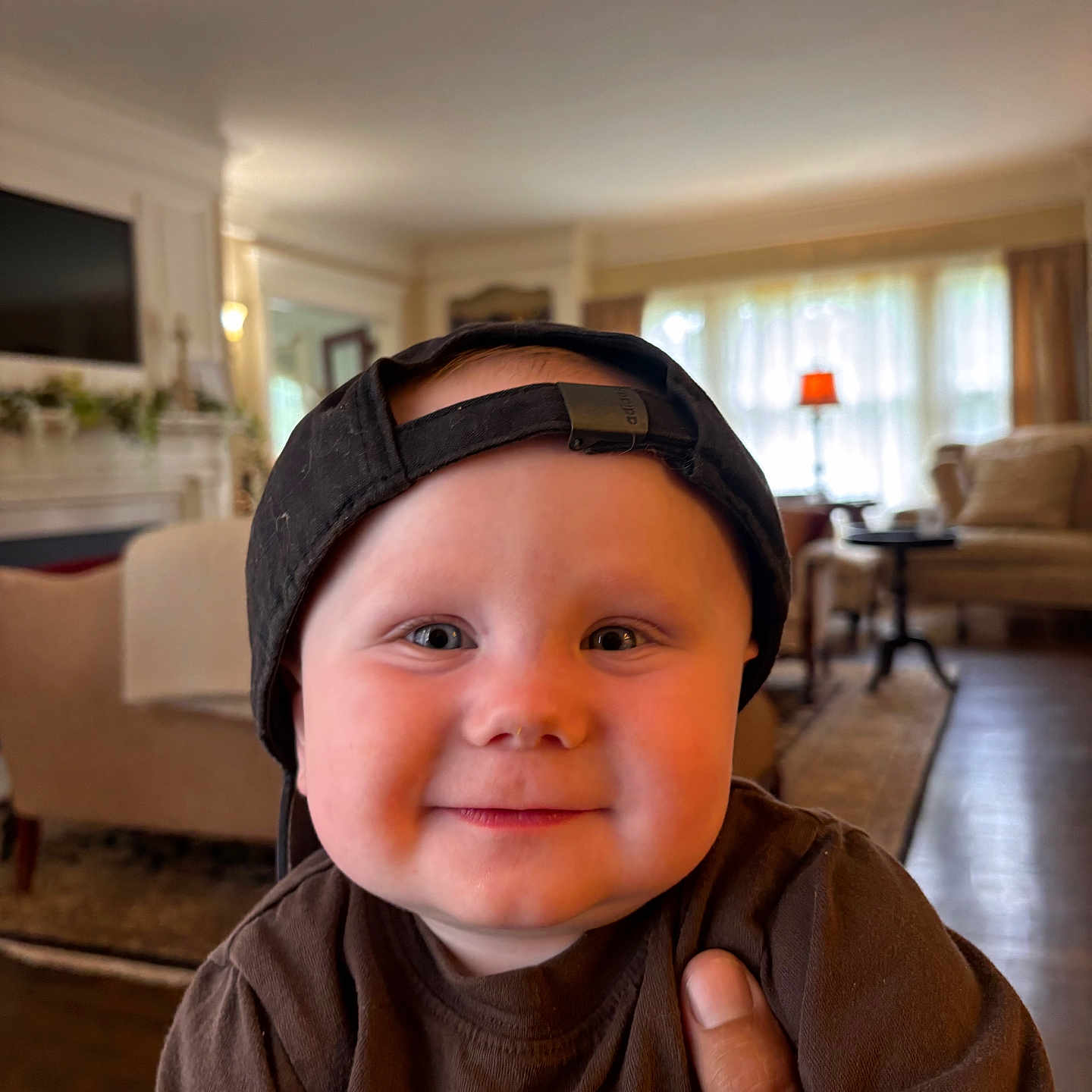 Canaan is registered to the contest to win money with this photo: baby, brown_shirt, cap, casual, cheeks, child, cozy, cute, face, furniture, hand, happy, headwear, home, indoor, living_room, person, portrait, smiling, toddler