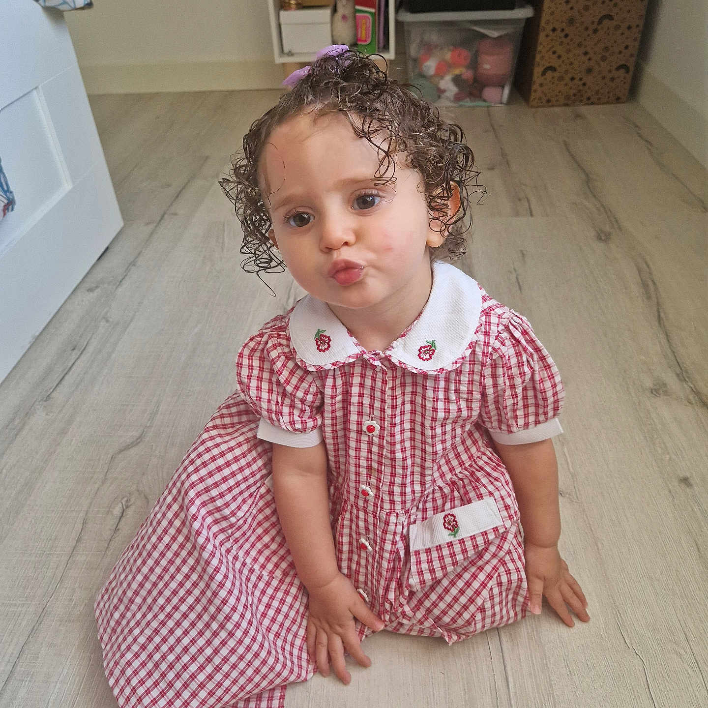 Eden a rejoint le concours — aidez-le/la à gagner de superbes lots ! baby, checkered, child, curly_hair, curly_hair_toddler, cute, dress, floor, foot, hand, indoor, person, pouting, red_and_white, room, sitting, storage, toddler, toy, wooden_floor