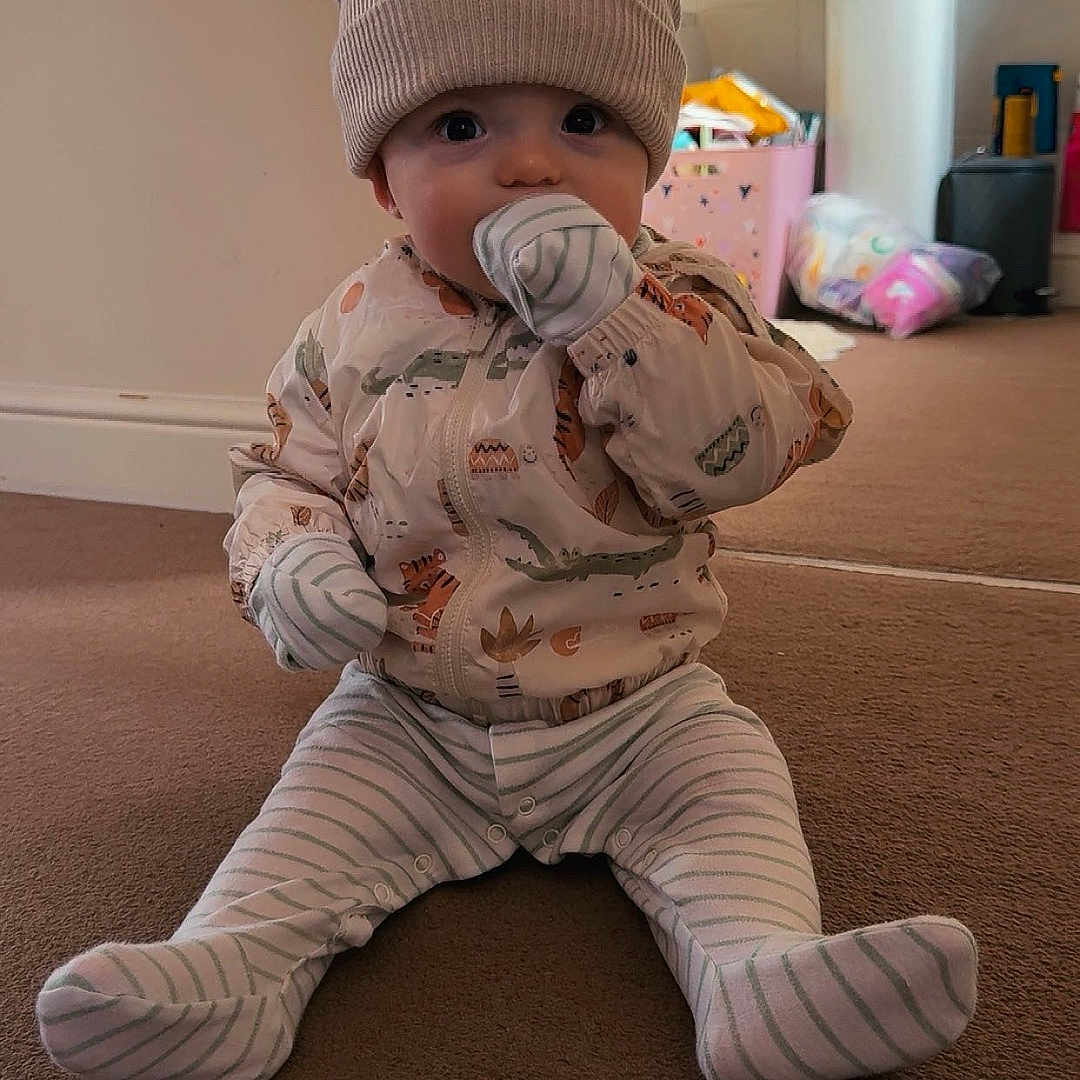 Elijah is registered to the contest to win money with this photo: baby, bathroom, carpet, child, clothing, cute, face, floor, hat, home, indoor, infant, mittens, pajamas, person, sink, sitting, striped_clothing, toy_box, young_child