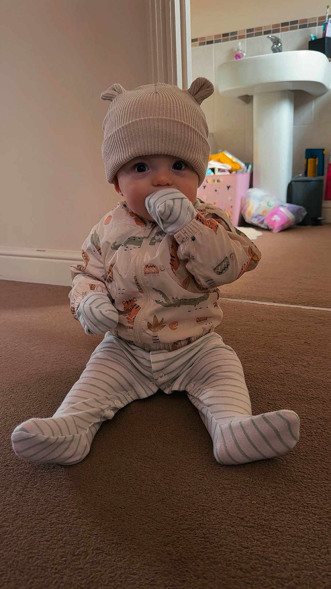 Elijah is registered to the contest to win money with this photo: baby, child, indoor, carpet, hat, mittens, striped_clothing, pajamas, sitting, floor, face, person, cute, home, bathroom, sink, toy_box, clothing, infant, young_child