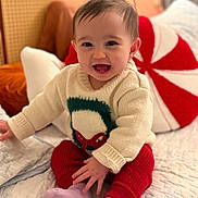 Violet is registered to the contest to win money with this photo: baby, child, smiling, sitting, sweater, red_pants, happy, indoors, bed, pillow, cozy, cute, portrait, knitwear, holiday, warm_lighting, infant, person, furniture, soft_texture