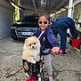 child, dog, puppy, bicycle, basket, sunglasses, person, car, audi, garage, outdoor, concrete, clothing, footwear, pet, happy, smile, toy, plant, casual