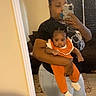 adult, baby, mirror_selfie, orange_clothing, casual, indoor, person, holding, jeans, black_shirt, sneakers, phone, reflection, carpet, doorframe, hairstyle, face, footwear, home_interior, expression