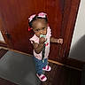 toddler, child, pacifier, pink_bows, pink_shoes, jeans, pink_shirt, wooden_door, door_handle, floor_mat, indoor, standing, curly_hair, hair_beads, expression, flooring, home, person, cute, young_child