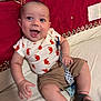 baby, child, clothing, couch, fabric, face, footwear, hand, happy, indoor, infant, pacifier, pattern, person, red, shirt, shorts, skin, smile, white