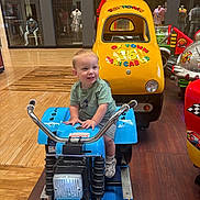 Carson joined the competition — help win amazing prizes! toddler, child, toy, motorcycle_ride, arcade, indoor, mall, smiling, yellow, blue, rubber_duck, taxi_cab, flooring, mannequin, clothing, footwear, play, ride, person, happy