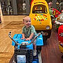 toddler, child, toy, motorcycle_ride, arcade, indoor, mall, smiling, yellow, blue, rubber_duck, taxi_cab, flooring, mannequin, clothing, footwear, play, ride, person, happy