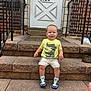 toddler, child, steps, stone, door, house_number, yellow_tshirt, shorts, sneakers, skateboard_graphic, railings, brick_wall, smiling, outdoor, front_door, person, young_child, casual_clothing, daylight, concrete