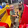 Carson is registered to the contest to win money with this photo: toddler, child, kiddie_ride, mall, shopping_mall, red, yellow, steering_wheel, toy_car, sneakers, shorts, green_shirt, smile, happy, indoor, floor, bench, teenager, mannequin, phone