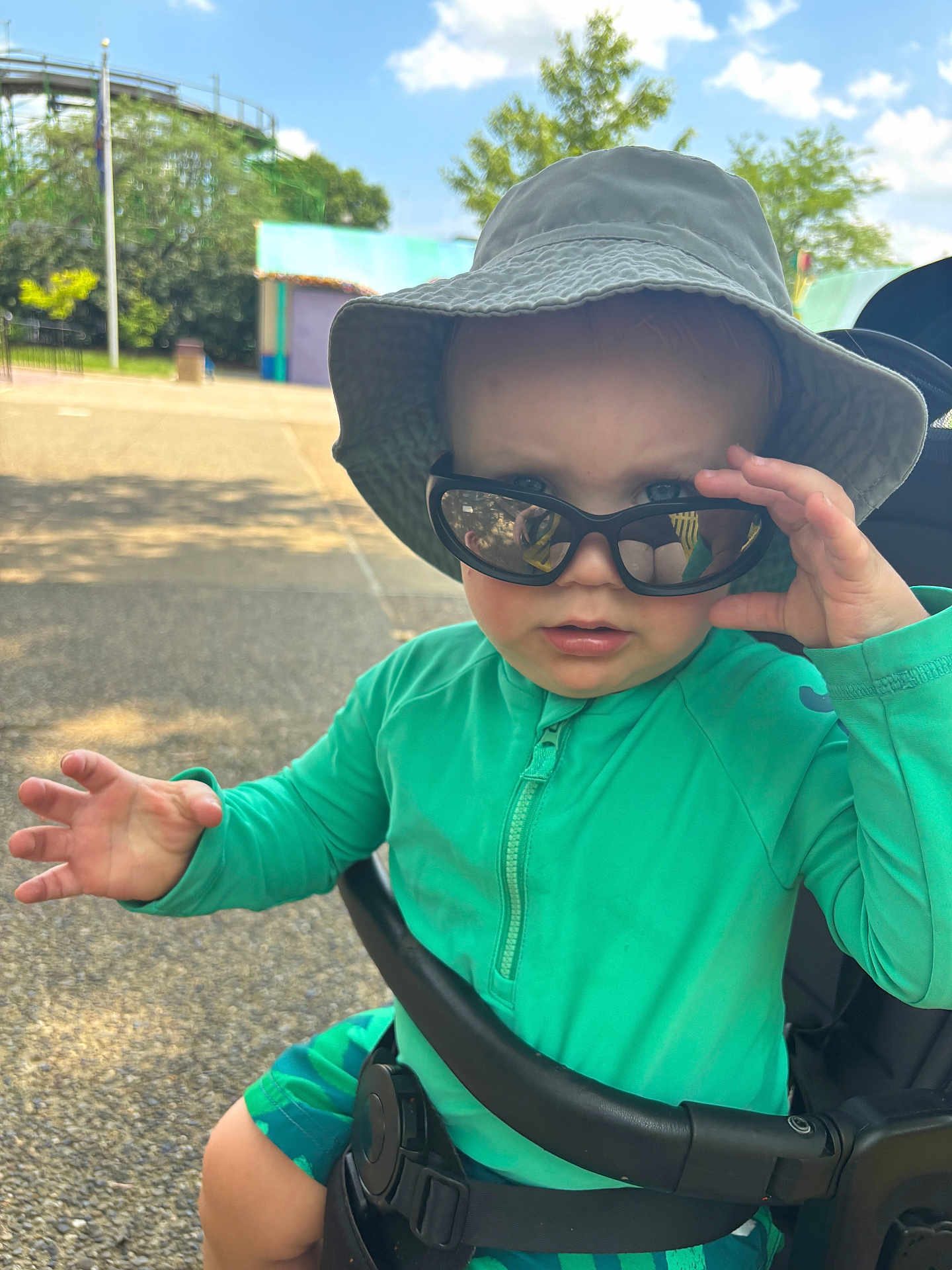 Carson joined the competition — help win amazing prizes! toddler, child, sunglasses, hat, stroller, outdoor, sunny, green_clothing, hand, face, expression, summer, park, seatbelt, blue_sky, trees, casual, cute, person, daytime