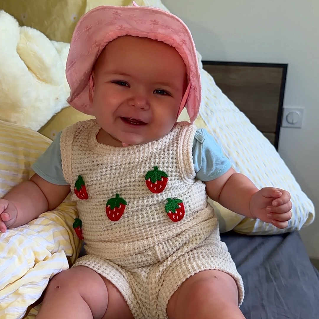 Thessa a rejoint le concours — aidez-le/la à gagner de superbes lots ! baby, child, pink_hat, knitted_romper, strawberry_pattern, smiling, sitting, bed, pillow, yellow_pillow, white_pillow, cute, happy, indoors, infant, legs, arms, face, headwear, cozy