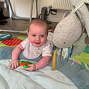 Albie is registered to the contest to win money with this photo: baby, infant, smiling, playmat, toy, indoor, floor, bib, cheeks, hand, colorful, soft_toy, baby_gear, wooden_floor, radiator, clothing, person, cute, happy, child