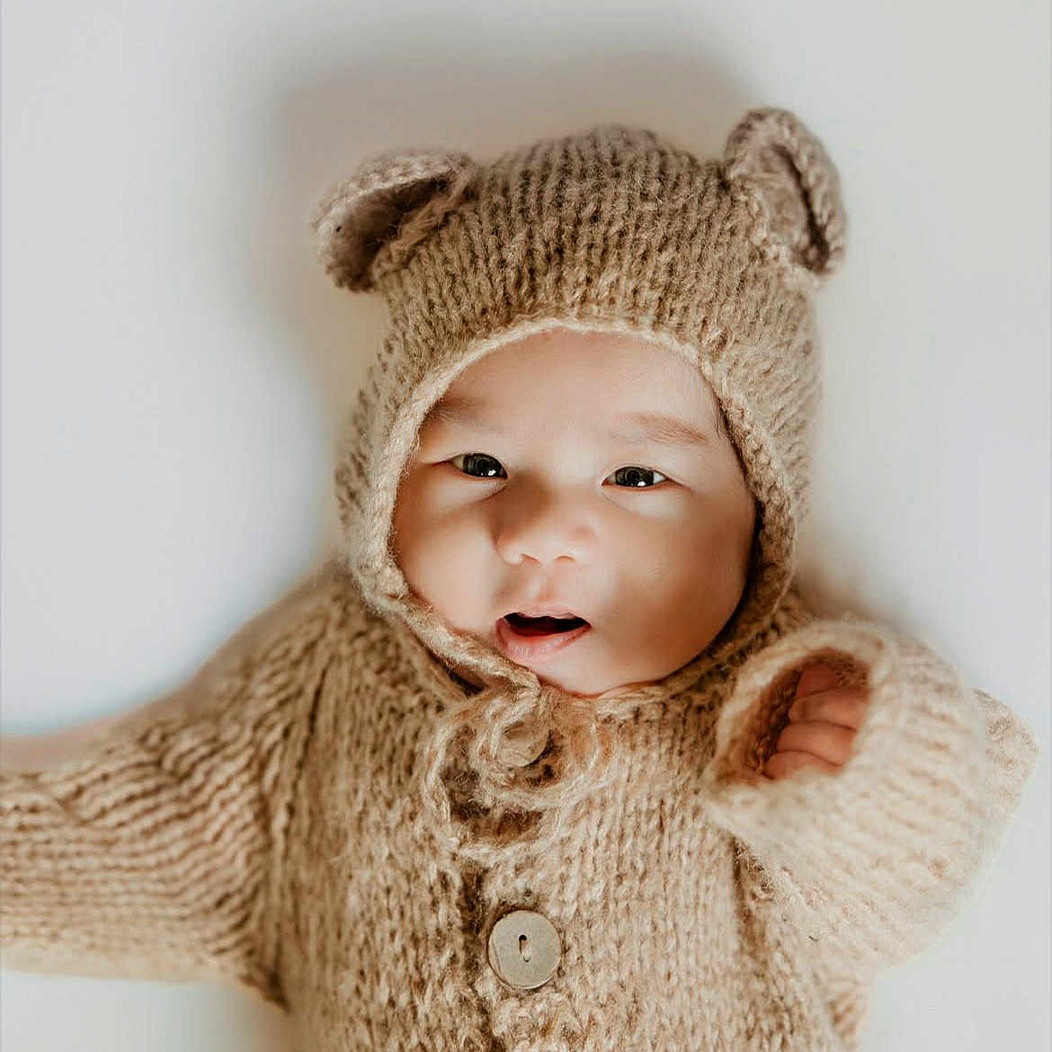 Erick is registered to the contest to win money with this photo: baby, infant, knitted_clothing, bear_ears, cute, portrait, child, warm, soft_texture, cozy, buttoned_sweater, hood, smiling, happy, indoor, closeup, face, small_hand, light_background, adorable