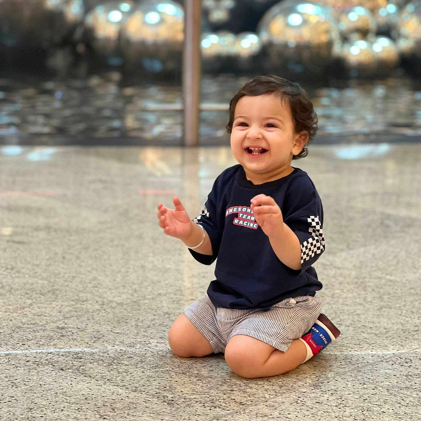 Viransh is registered to the contest to win money with this photo: bright_lighting, casual_clothing, cheerful, child, curly_hair, cute, floor, happy, indoor, kneeling, metallic_spheres, navy_shirt, person, playful, portrait, reflection, smiling, striped_shorts, toddler, young_child