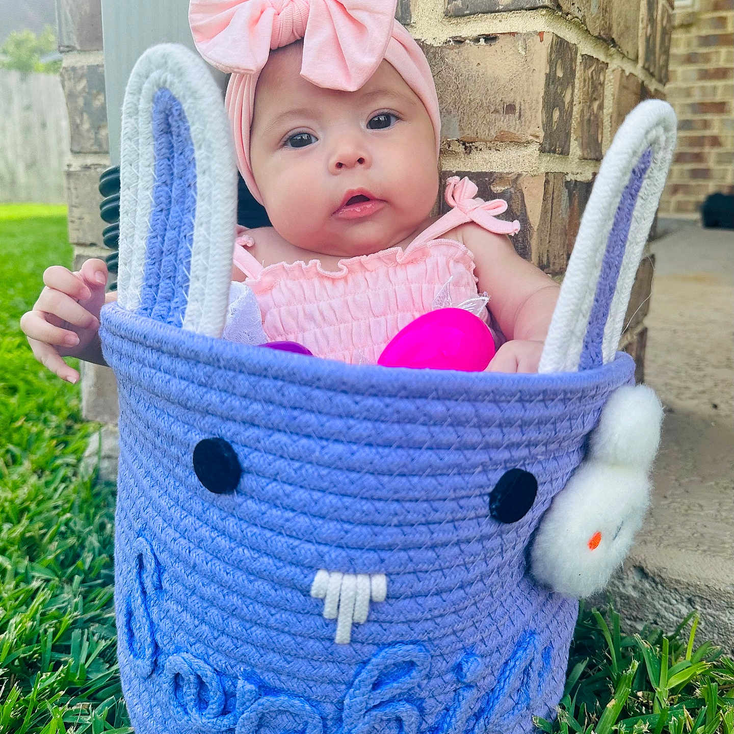 Sophia is registered to the contest to win money with this photo: baby, basket, brick_wall, bunny_ears, child, clothing, cute, face, grass, hands, headband, infant, outdoor, person, pink_bow, portrait, purple, smile, soft_light, toy