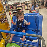 Emmett is registered to the contest to win money with this photo: toddler, child, shopping_cart, store, snack, blue_seat, person, aisle, grocery, food, packaging, curly_hair, clothing, floor, shelf, shopping, retail, supermarket, smile, indoor