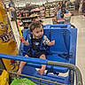 toddler, child, shopping_cart, store, snack, blue_seat, person, aisle, grocery, food, packaging, curly_hair, clothing, floor, shelf, shopping, retail, supermarket, smile, indoor