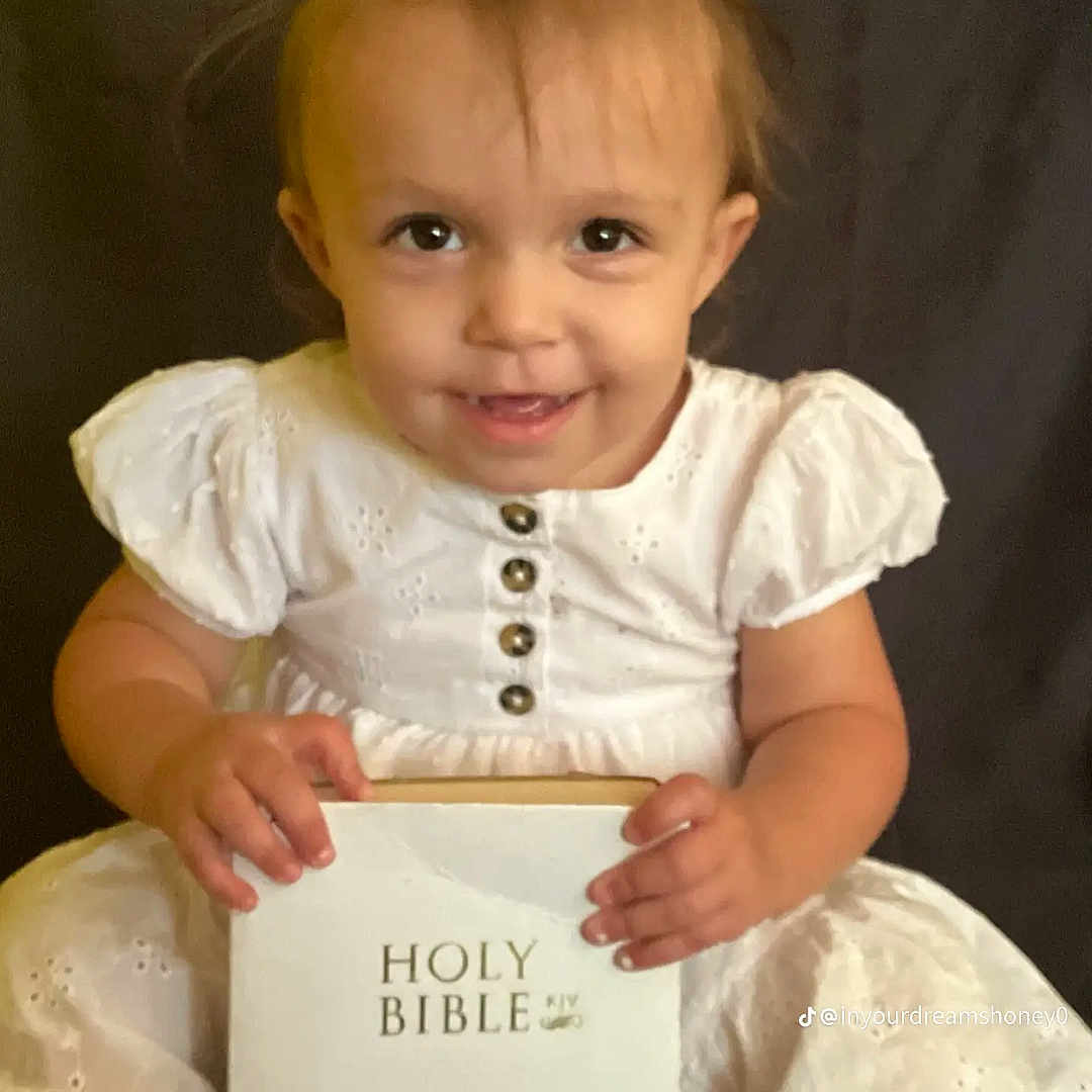 Catherine Lawson is registered to the contest to win money with this photo: bible, black_background, book, buttoned_dress, child, cute, face, hands, happy, holding_object, indoor, innocence, person, pigtails, portrait, sitting, smiling, toddler, white_dress, young_child