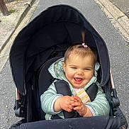 Lina participe au concours pour gagner de l'argent avec cette photo : baby, child, stroller, happy, smiling, outdoor, jacket, clapping, person, infant, pony_tail, road, sidewalk, black_stroller, cute, joyful, seated, winter_clothing, young_child, daytime