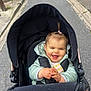 baby, child, stroller, happy, smiling, outdoor, jacket, clapping, person, infant, pony_tail, road, sidewalk, black_stroller, cute, joyful, seated, winter_clothing, young_child, daytime