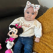 Victoire participe au concours pour gagner de l'argent avec cette photo : baby, child, blue_eyes, headband, floral, sweater, text_on_clothing, plush_toy, minnie_mouse, couch, cushion, blanket, jeans, sitting, indoor, cute, portrait, person, soft_lighting, comfort