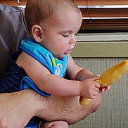 Joseph is registered to the contest to win money with this photo: arm, baby, bib, child, clothing, curious, face, head, holding, human, indoor, infant, light, person, portrait, sitting, skin, table, wooden_blinds, yellow_object
