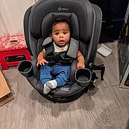 Zayne is registered to the contest to win money with this photo: baby, child, infant, car_seat, sitting, wide_eyed, indoors, portrait, safety_harness, blue_pants, socks, cup_holder, floor, home, furniture, appliance, laundry, red_stool, photography, surprised