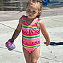 child, girl, swimsuit, watermelon_pattern, water_shoe, outdoor, splash_pad, concrete, sunny, hair_buns, playground, summer, water_play, footwear, walking, preschooler, casual, daylight, cute, toddler