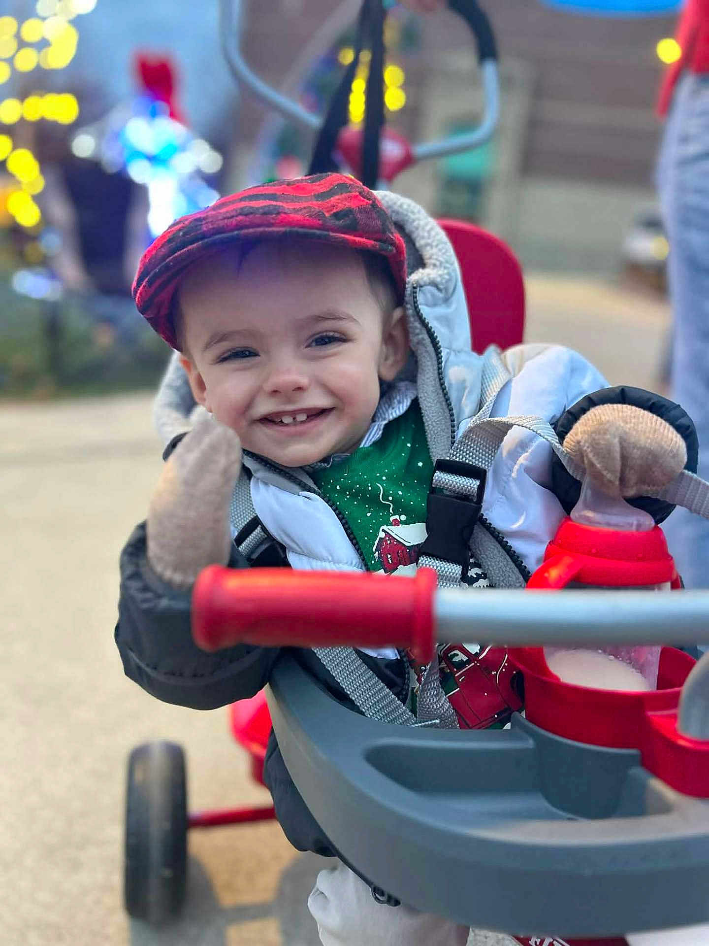 Brody Dean is registered to the contest to win money with this photo: toddler, child, smiling, hat, jacket, mittens, stroller, toy, outdoor, winter_clothing, happy, face, portrait, holiday_lights, blurred_background, red, green, cozy, cute, waving