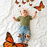 Anna is registered to the contest to win money with this photo: baby, infant, butterflies, green_shirt, denim_shorts, ripped_jeans, headband, white_bedspread, lying_down, orange_butterflies, cute, playful, child, soft_lighting, indoor, adorable, portrait, happy, nature_theme, cozy
