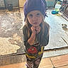 child, girl, purple_wig, mickey_mouse, shirt, leggings, socks, indoor, floor, cracked_floor, worn_floor, person, toy, window, door, bucket, shelf, food_cans, water_dispenser, expression