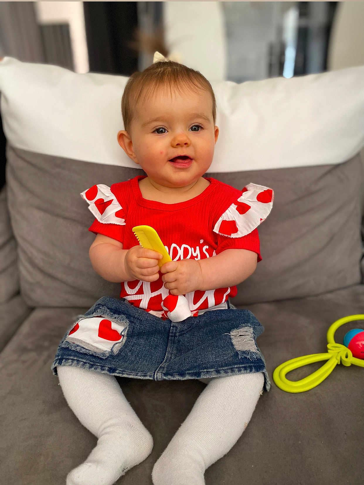 Noëlya a rejoint le concours — aidez-le/la à gagner de superbes lots ! baby, child, smiling, red_shirt, denim_skirt, white_tights, comb, toy, rattle, couch, indoor, happy, sitting, infant, cute, heart_pattern, clothing, person, portrait, home