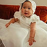 baby, child, dress, white_dress, lace_bonnet, couch, indoor, portrait, cute, adorable, smiling, infant, soft_lighting, fabric, sitting, young_child, chubby_arms, face, person, homedecor