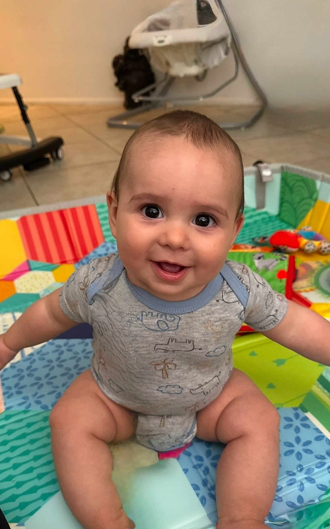 Theo joined the competition — help win amazing prizes! baby, infant, smiling, face, big_eyes, onesie, play_mat, toys, tile_floor, baby_swing, sitting, chubby_cheeks, diaper, happy_expression, portrait, indoor, colorful, cute, arms_out, playtime