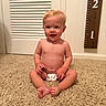 baby, child, diaper, carpet, smile, sitting, indoor, measurement_board, growth_chart, blond_hair, happy, cute, skin, person, floor, room, young_child, infant, head, face
