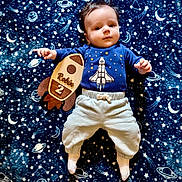 Robin is registered to the contest to win money with this photo: baby, infant, child, blanket, space_theme, rocket, wooden_sign, blue_shirt, pants, socks, lying_down, stars, planets, moon, cute, portrait, indoors, young_child, comfortable, adorable